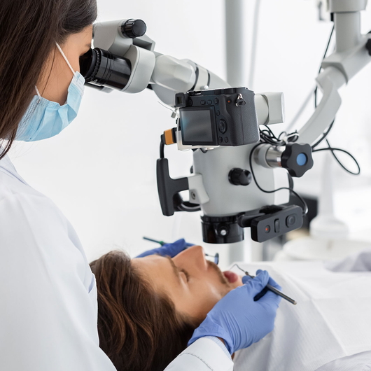 Eine Zahnärztin mit Mundschutz und Handschuhen benutzt ein Dentalmikroskop, während er einen Patienten behandelt, der in einer modernen Zahnklinik auf einem Stuhl liegt.