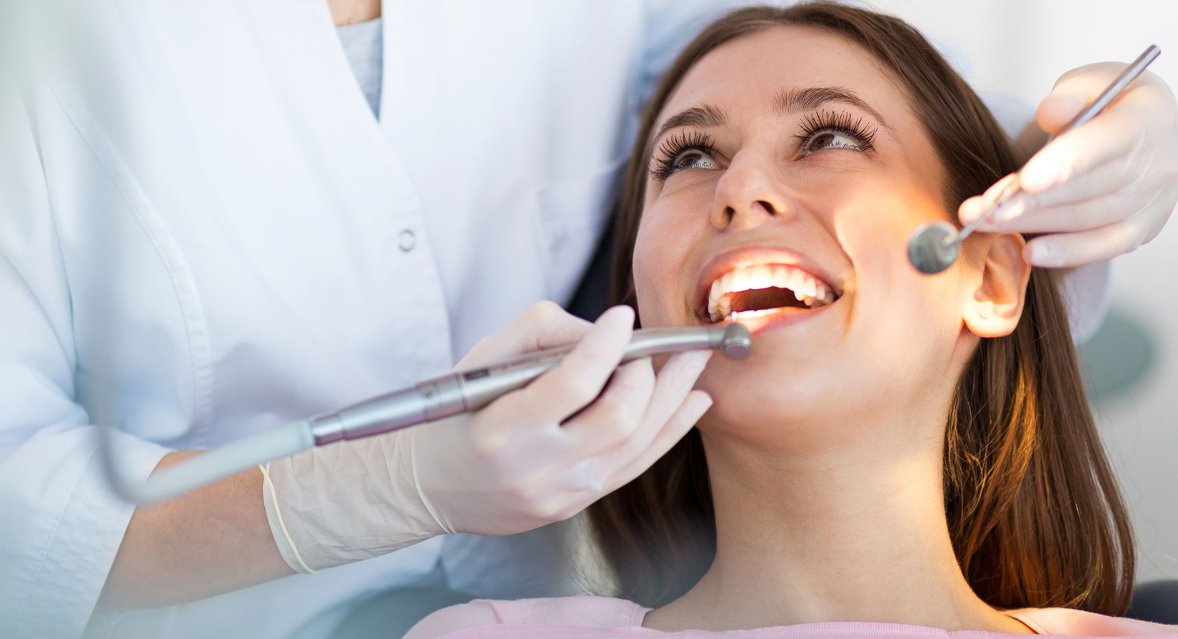 Eine Frau sitzt in einem Zahnarztstuhl und lächelt mit offenem Mund, während eine Zahnärztin mit weißen Handschuhen während einer zahnärztlichen Untersuchung Instrumente an ihre Zähne hält.