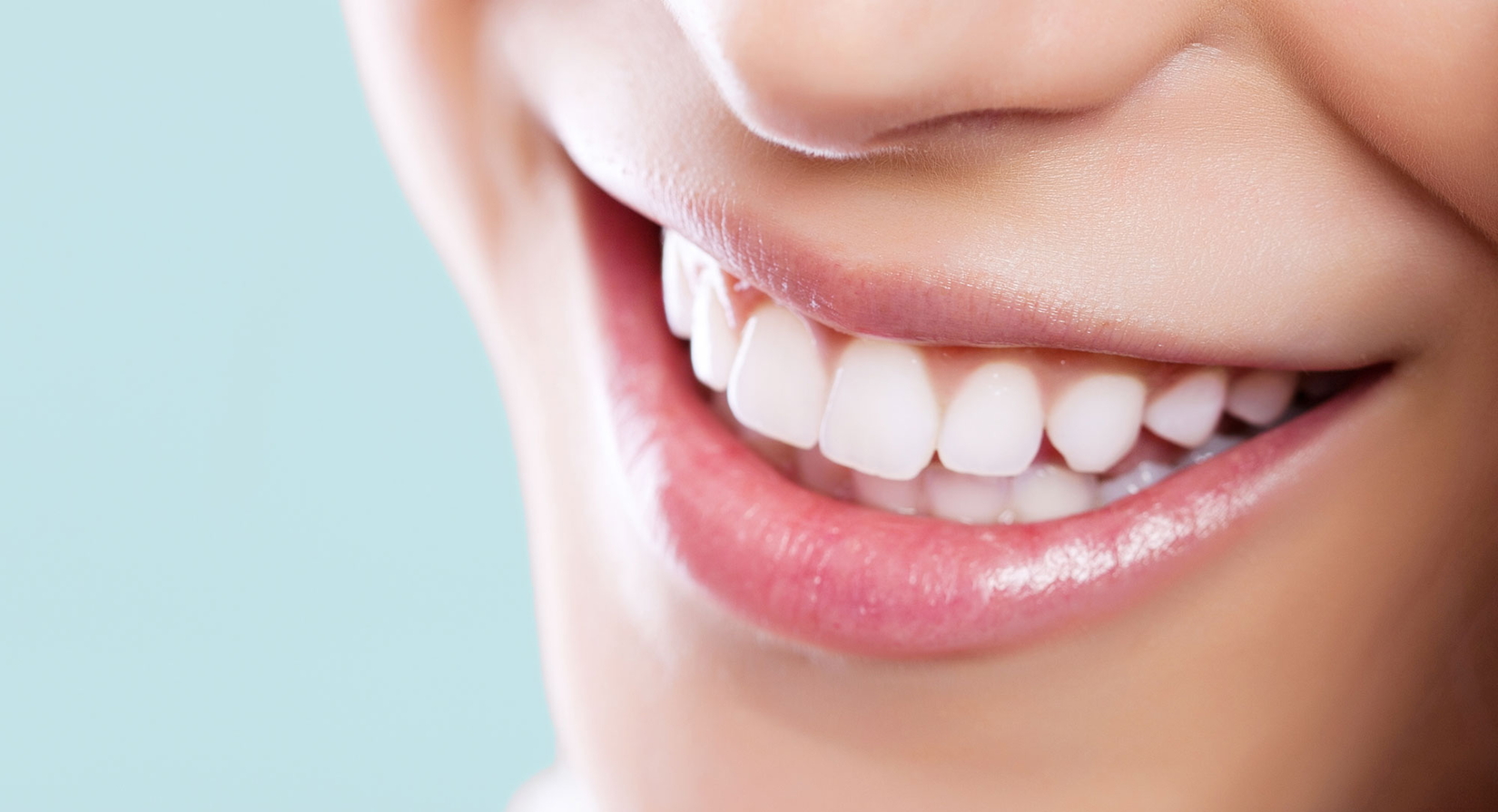 Nahaufnahme des Mundes einer lächelnden Person mit geraden weißen Zähnen und rosafarbenen Lippen, wobei glatte Haut und ein Teil der Nase sichtbar sind. Der Hintergrund ist ein sanftes Blau.
