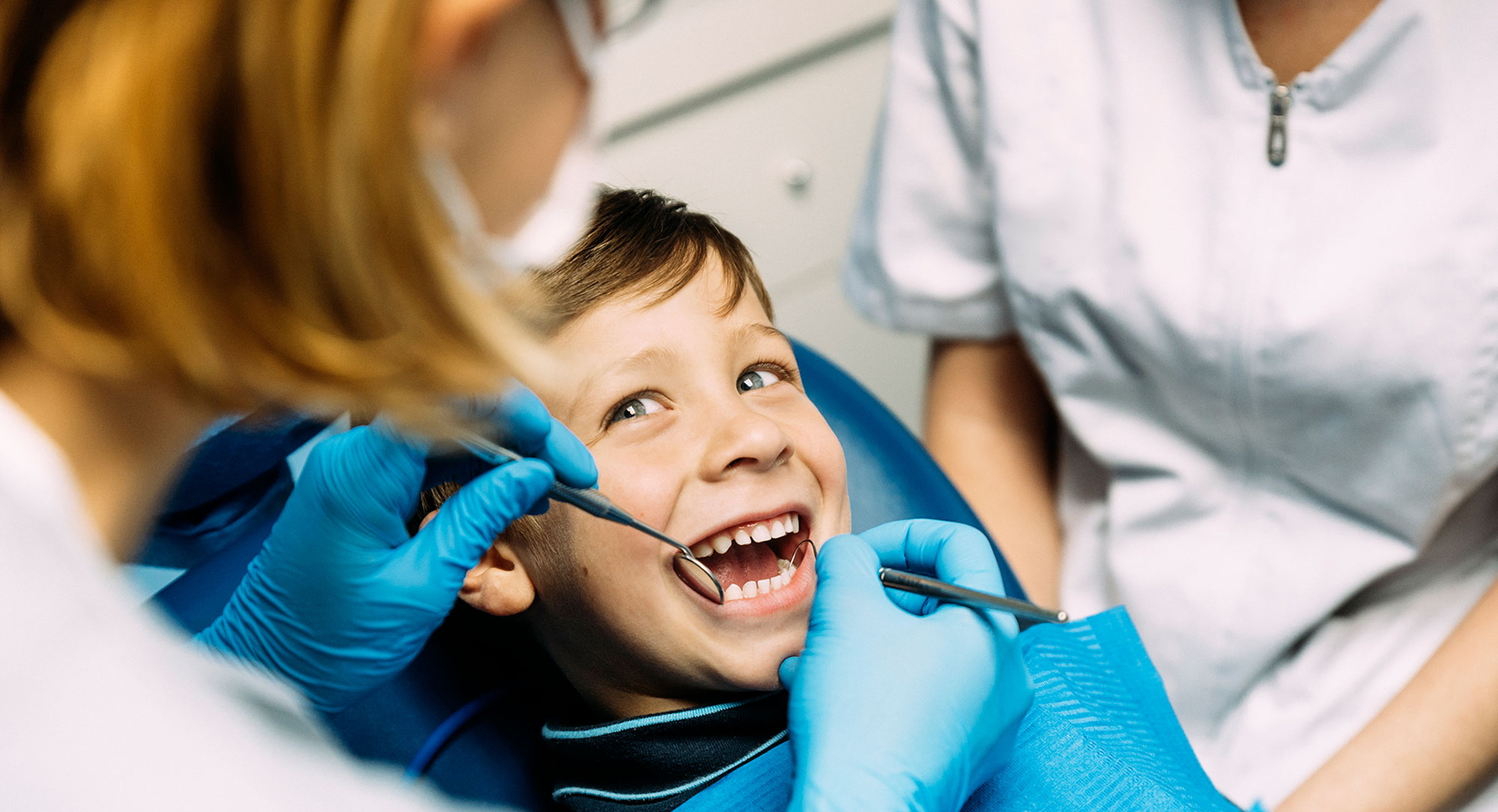 Ein kleiner Junge sitzt lächelnd in einem Zahnarztstuhl, während eine Zahnärztin mit blauen Handschuhen seine Zähne mit zahnmedizinischen Instrumenten untersucht. Eine weitere Person in einem weißen Kittel sitzt daneben.