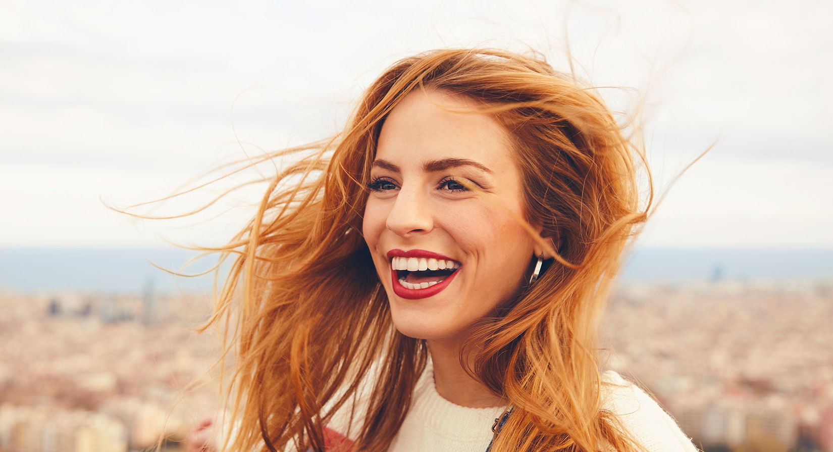 Eine junge Frau mit langem, vom Wind zerzaustem rotem Haar lächelt im Freien, mit einer verschwommenen Stadtlandschaft und einem bewölkten Himmel im Hintergrund. Sie trägt einen leichten Pullover und Ohrringe.